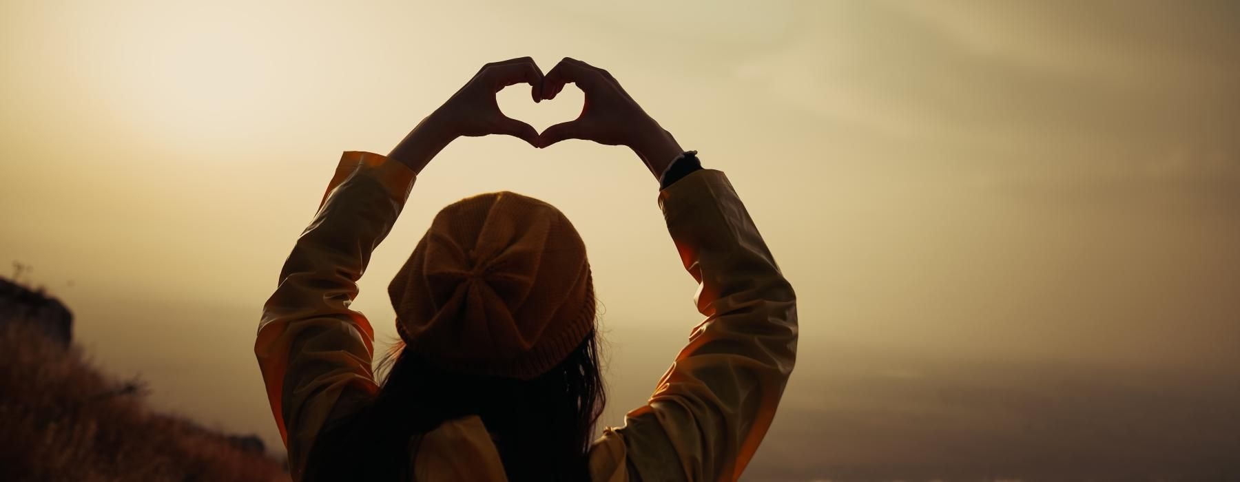 woman stands on a mountain top with her hands up in the air forming a heart