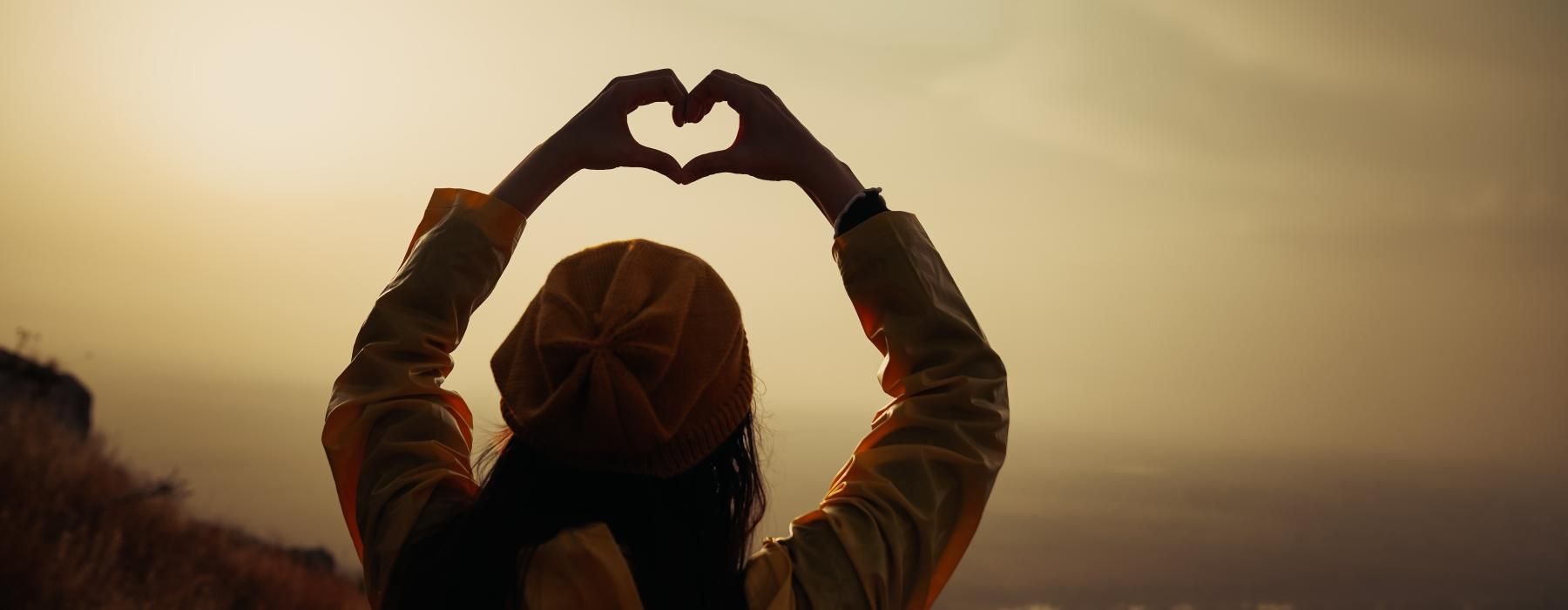 woman stands on a mountain top with her hands up in the air forming a heart